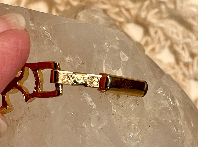 May include: A gold-toned metal bracelet with a clasp that reads "AVON" in all caps.