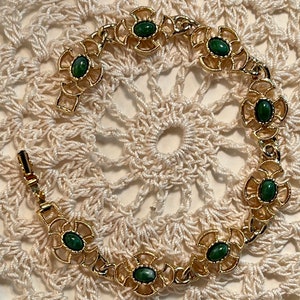 May include: A gold bracelet with green stones set in a floral design. The bracelet is on a white crocheted surface.
