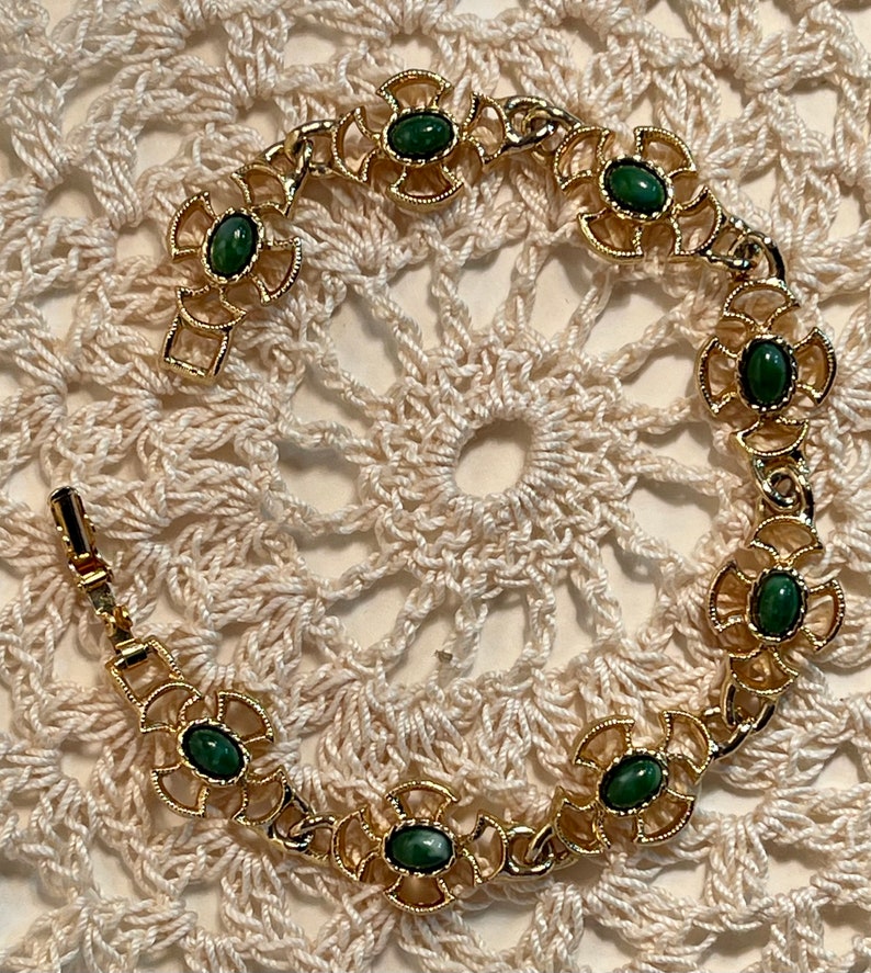 May include: A gold bracelet with green stones set in a floral design. The bracelet is on a white crocheted surface.