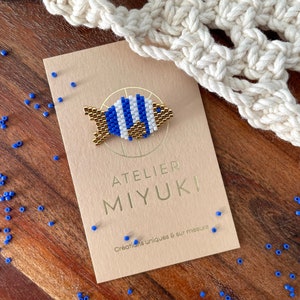 Puede incluir: Un broche de pez con cuentas azules y blancas y detalles dorados descansa sobre una tarjeta beige. La tarjeta dice "ATELIER MIYUKI" y "Créations uniques & sur mesure". Hay cuentas azules dispersas sobre una superficie de madera.