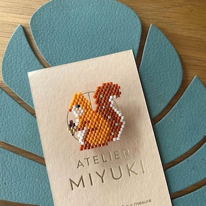 May include: A beaded squirrel pin in shades of orange, white, and brown, set against a tan card with the text "ATELIER MIYUKI". The pin is a detailed, handcrafted accessory, perfect for adding a touch of nature-inspired charm to any outfit.