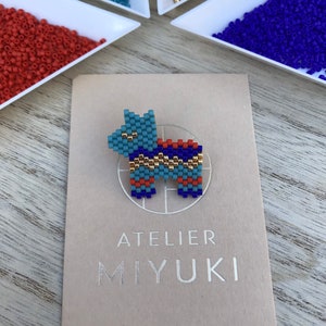 Peut inclure: Épingle à nourrice en forme de chien, composée de perles bleues claires, orange, dorées et bleu foncé. L'épingle est présentée sur une carte avec le texte "ATELIER MIYUKI" et "Bijoux uniques & sur mesure". Des perles de différentes couleurs sont en arrière-plan.