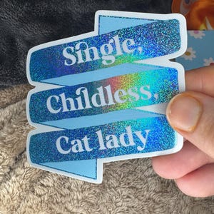 May include: A blue sticker with the words "Single, Childless, Cat lady" in white text. The sticker has a holographic, glittery effect. The sticker is held by a person's hand.