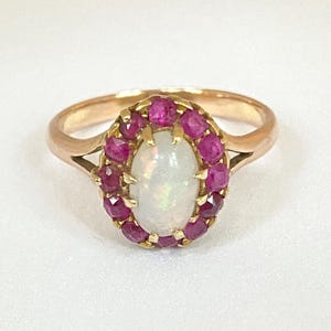 Può includere: Un anello d'oro con una pietra centrale ovale di opale iridescente, circondata da un'aureola di piccole gemme rosse. La fascia dell'anello è in oro lucido e l'incastonatura è delicata e ornata. Un gioiello classico.