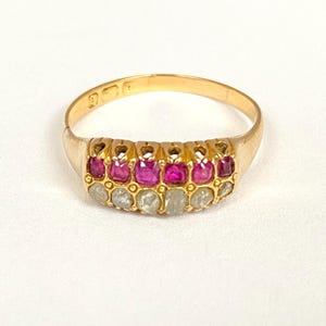 May include: A gold ring featuring a row of alternating red and clear gemstones. The ring has a classic design with a polished gold band. The gemstones are set closely together, creating a continuous line of color and sparkle. The ring is displayed on a white background.