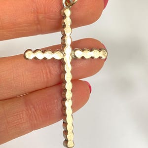 May include: A gold-colored cross pendant with a textured, scalloped design. The cross has a small loop at the top for hanging. The pendant is held against a light background.