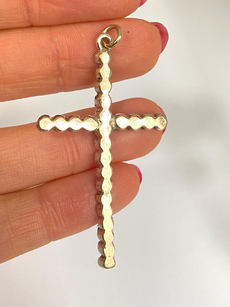May include: A gold-colored cross pendant with a textured, scalloped design. The cross has a small loop at the top for hanging. The pendant is held against a light background.