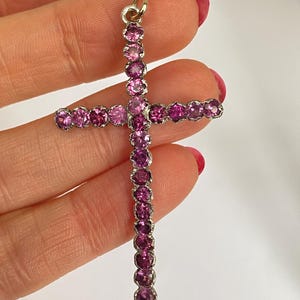 May include: A gold-toned cross pendant adorned with numerous pink gemstones. The gemstones are set closely together, creating a continuous line of color. The cross has a loop at the top for a chain.