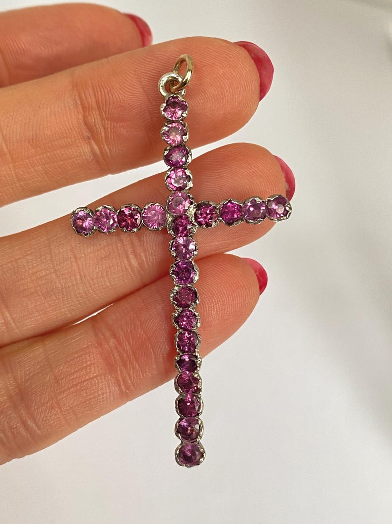 May include: A gold-toned cross pendant adorned with numerous pink gemstones. The gemstones are set closely together, creating a continuous line of color. The cross has a loop at the top for a chain.