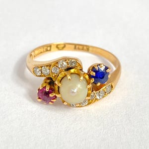 May include: A gold ring featuring a central pearl, a ruby, and a sapphire, accented with small diamonds. The ring has a delicate, ornate design with a polished finish. The gemstones are set in a cluster arrangement.