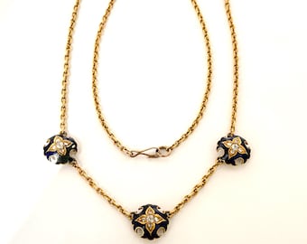 Puede incluir: Un collar dorado con tres cuentas esmaltadas en azul oscuro y blanco, cada una con un diseño floral en forma de diamante. El collar tiene una cadena fina y un cierre de gancho.