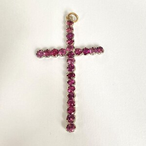 May include: A pendant in the shape of a cross, adorned with numerous pink gemstones. The cross features a gold-colored loop at the top for hanging. The gemstones are set in a silver-colored metal.