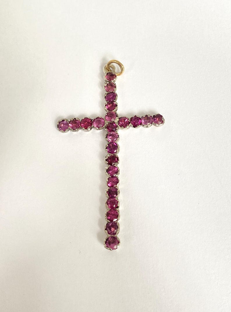 May include: A pendant in the shape of a cross, adorned with numerous pink gemstones. The cross features a gold-colored loop at the top for hanging. The gemstones are set in a silver-colored metal.