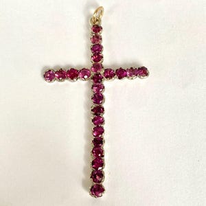 May include: A pendant in the shape of a cross, adorned with numerous round, deep pink gemstones. The gemstones are set in a silver-colored metal, and the top has a small gold-colored loop for a chain.
