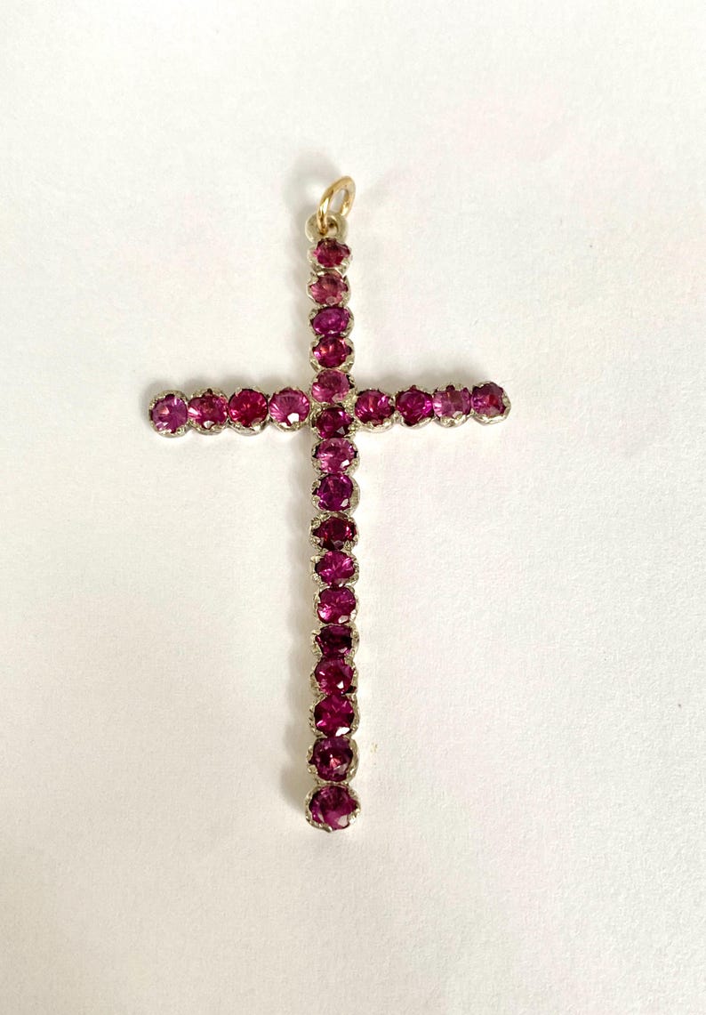 May include: A pendant in the shape of a cross, adorned with numerous round, deep pink gemstones. The gemstones are set in a silver-colored metal, and the top has a small gold-colored loop for a chain.