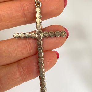 May include: Silver-toned cross pendant with a loop at the top for a chain. The cross is adorned with a series of small, circular embellishments along its arms and vertical bar. The pendant is held against a white background.