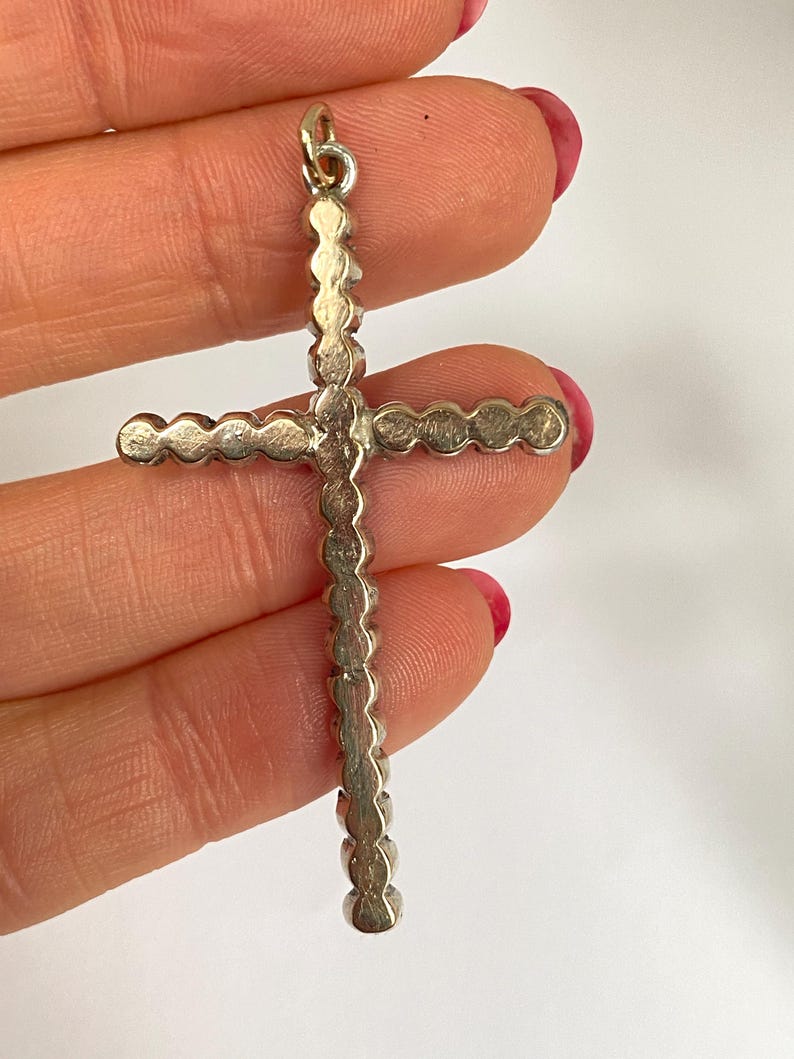 May include: Silver-toned cross pendant with a loop at the top for a chain. The cross is adorned with a series of small, circular embellishments along its arms and vertical bar. The pendant is held against a white background.