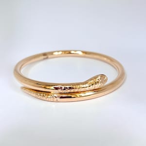 Puede incluir: Un brazalete dorado con forma de serpiente. El brazalete se enrolla, con la cabeza y la cola de la serpiente superpuestas. La cabeza y la cola de la serpiente tienen detalles texturizados. El brazalete está sobre un fondo blanco.