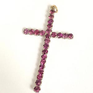 May include: A pendant in the shape of a cross, featuring a series of pink gemstones set in a silver-colored metal. The top of the cross has a small gold-colored loop for a chain. The gemstones are a vibrant shade of pink.