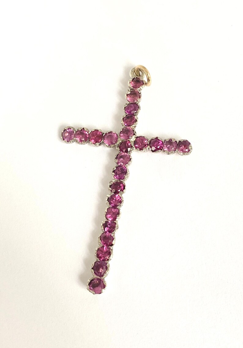 May include: A pendant in the shape of a cross, featuring a series of pink gemstones set in a silver-colored metal. The top of the cross has a small gold-colored loop for a chain. The gemstones are a vibrant shade of pink.