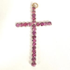 May include: A cross-shaped pendant featuring numerous round, pink gemstones set in a silver-colored metal. The pendant has a gold-colored loop at the top for hanging. The gemstones are closely set, creating a continuous line of color.