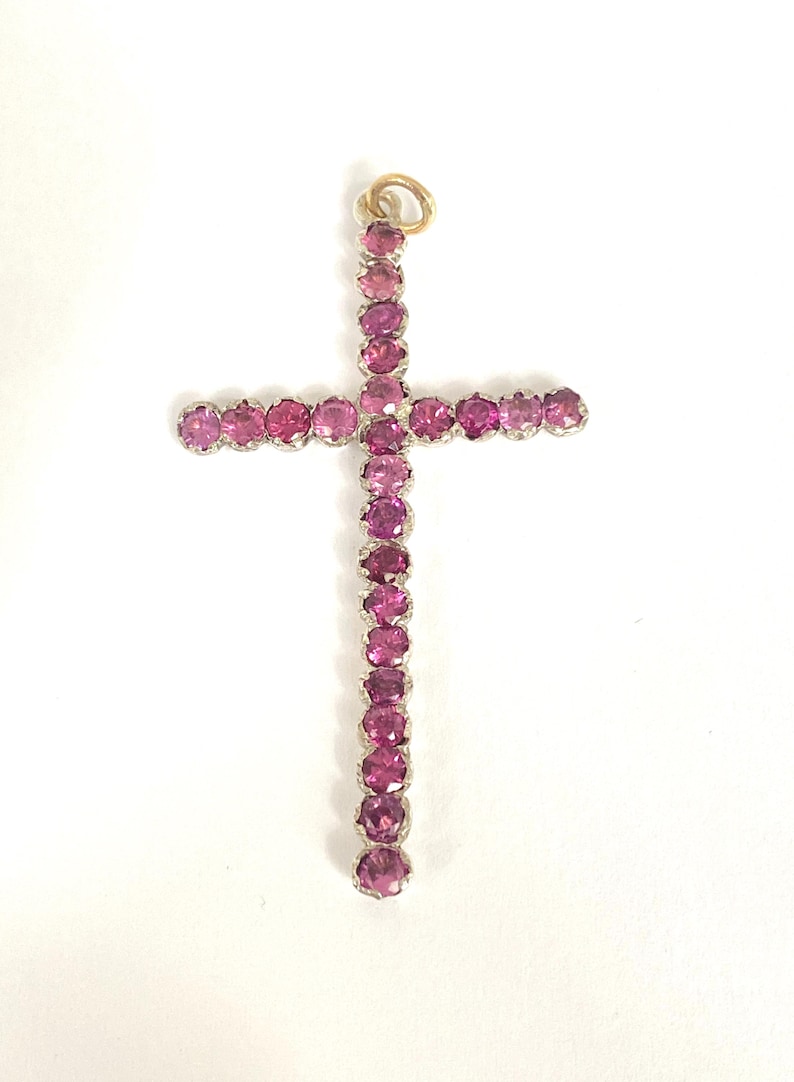 May include: A cross-shaped pendant featuring numerous round, pink gemstones set in a silver-colored metal. The pendant has a gold-colored loop at the top for hanging. The gemstones are closely set, creating a continuous line of color.
