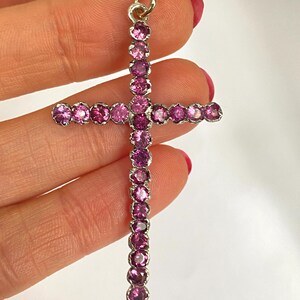 May include: A silver cross pendant adorned with numerous round, pink gemstones. The gemstones are set closely together, creating a continuous line of color. The cross has a small loop at the top for a chain.