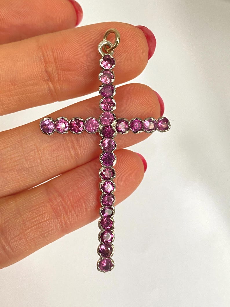 May include: A silver cross pendant adorned with numerous round, pink gemstones. The gemstones are set closely together, creating a continuous line of color. The cross has a small loop at the top for a chain.