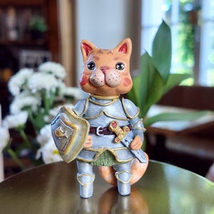 May include: A small figurine of an orange tabby cat dressed in silver knight armour. The cat holds a shield with a gold fish emblem and a sword at its waist. The armour features gold trim, and the cat has large dark eyes, pink cheeks, and a green tunic.