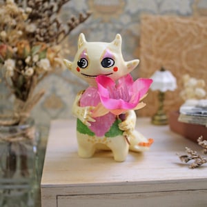 May include: A whimsical, hand-painted ceramic figurine of a creature holding a pink flower. The figurine has a cream-colored body with black eyes, pink accents, and a green and pink floral design. The creature has small horns and a tail.