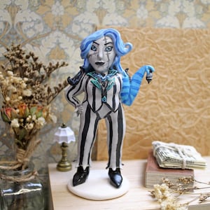 May include: A handmade figurine of a character with blue hair and a black and white striped suit. The figure is standing on a white base, with black shoes. The character has a blue striped arm and is posed with one hand on their hip.