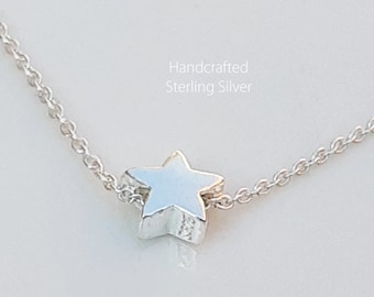 May include: A handcrafted sterling silver necklace featuring a small, three-dimensional star pendant. The star is silver and has a light blue hue. The chain is delicate and also silver. The text "Handcrafted Sterling Silver" is visible.