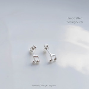 May include: A pair of handcrafted sterling silver stud earrings in the shape of musical notes. The earrings are displayed on a white surface. The text "Handcrafted Sterling Silver" is visible. The Etsy shop name is also visible.
