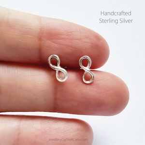 Può includere: Un paio di orecchini in argento sterling a forma di simbolo di infinito. Gli orecchini sono realizzati a mano e sono mostrati sul dito di una persona.