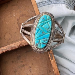 May include: A silver cuff bracelet with a large, oval turquoise stone inlay. The turquoise is a bright blue color and is cut into geometric shapes. The bracelet has a detailed, ornate design.