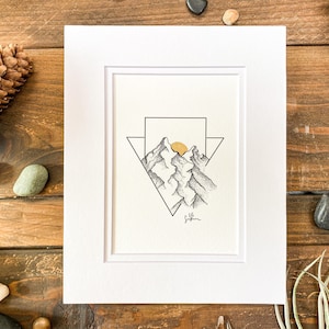 May include: A black and white line drawing of mountains with a gold sun behind them. The drawing is framed in a white mat and a white frame.