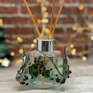 May include: A clear glass diffuser bottle with a silver cap and wooden reeds. The glass holder is decorated with green and red glass beads and a green leaf design. The background includes a brick wall and fairy lights.