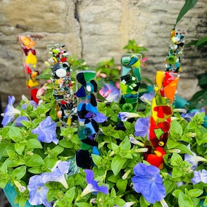 May include: Colourful glass garden stakes made of mosaic glass pieces. The stakes are arranged in a flower pot with purple flowers.