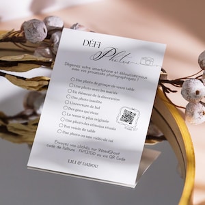 Peut inclure: Une carte blanche avec le texte "DÉFI Photos" et une liste de défis photo, avec un code QR. La carte est à côté d'une branche avec des baies décoratives blanches et marron. L'arrière-plan est de couleur neutre floue.