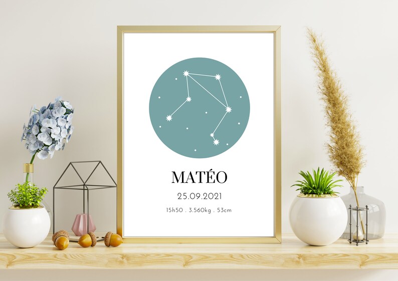 May include: A framed print featuring a teal circle with a white Libra constellation design. Below the circle is the name "MAT&Eacute;O", followed by the date, time, weight, and height. The print is displayed on a wooden shelf with decorative plants.