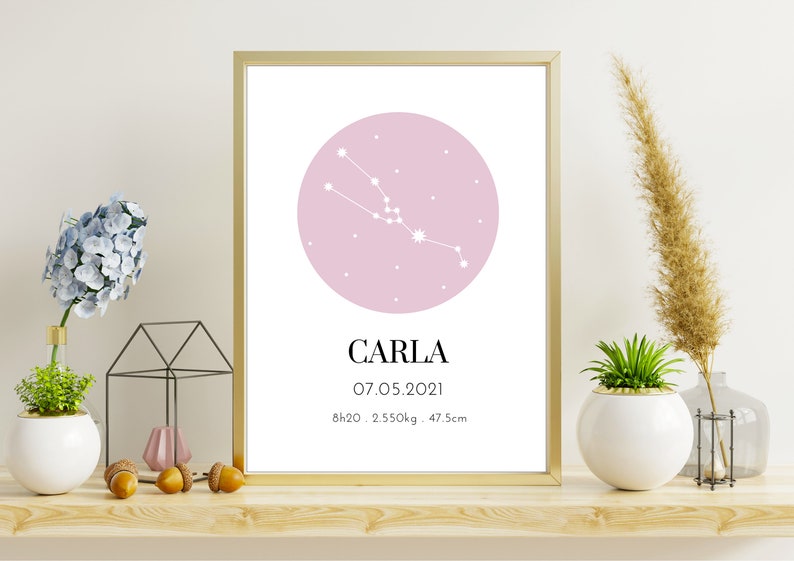 May include: A framed print featuring a pink circle with a white constellation design. The name "CARLA" is below the circle, along with the date 07.05.2021, and measurements: 8h20, 2.550kg, and 47.5cm. The frame is gold.