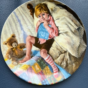 Könnte beinhalten: Dekorative Platte mit einer detaillierten Illustration eines Kindes, das ein Buch mit dem Titel "Bedtime Stories" hält. Das Kind sitzt auf einem Patchwork-Quilt, mit einem Teddybären. Die Platte hat eine farbenfrohe Illustration.
