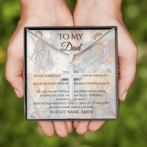 Könnte beinhalten: Eine silberne Kreuz-Anhänger-Halskette in einer Geschenkbox. Die Box trägt den Text "TO MY Dad" und eine religiöse Botschaft. Die Halskette wird in offenen Händen vor einem verschwommenen Hintergrund gehalten.