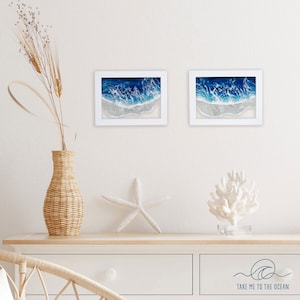 May include: Two framed ocean wave paintings with white frames and blue and white resin art. The paintings are hanging on a white wall above a white dresser with a wicker vase and a white starfish.