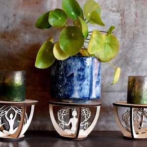 May include: Three wooden candle holders featuring a cut-out design of a person meditating in a tree pose. The holders are displayed against a grey backdrop with a potted plant in the centre.