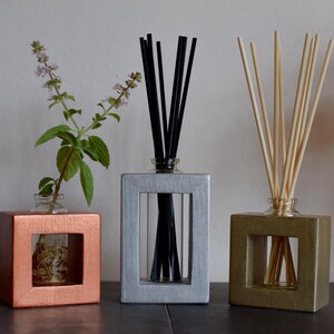 May include: Three square wooden diffusers with glass bottles and reeds. The diffusers are painted in copper, silver, and green. The copper diffuser has a small plant inside.