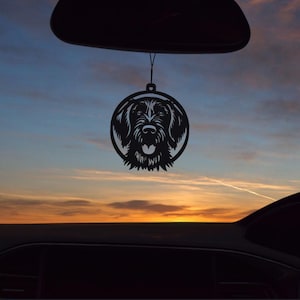 May include: Black metal dog silhouette car charm. The charm features a detailed cutout of a dog's face within a circular frame. The charm hangs from a black string, suspended against a sunset sky.