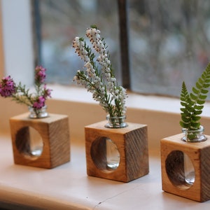 May include: Three small wooden cube vases with clear glass cylinders inside, each holding a different type of flower. The vases are arranged in a row on a white windowsill.