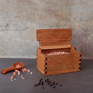 May include: A wooden salt box with a hinged lid, filled with pink Himalayan salt. A small wooden spoon and scattered salt crystals are next to the box. Black peppercorns are also visible. The box is made of wood.