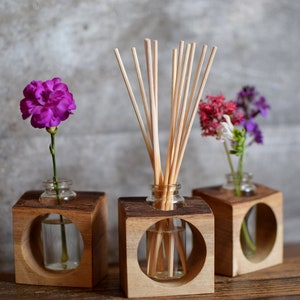 May include: Three wooden cube-shaped vases with round openings, each holding a different type of flower arrangement. The vase on the left holds a single purple carnation, the middle vase holds a diffuser with wooden sticks, and the right vase holds a bouquet of purple and white flowers.